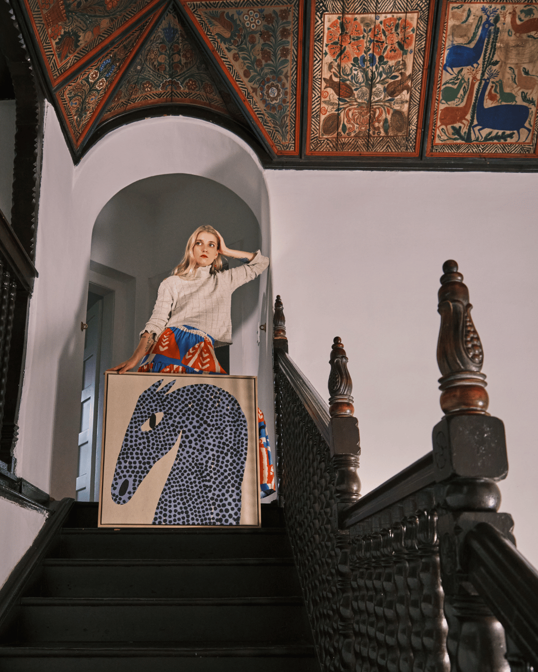 Woman standing holding artwork on staircase with hand painted floral motif decorative ceiling 
