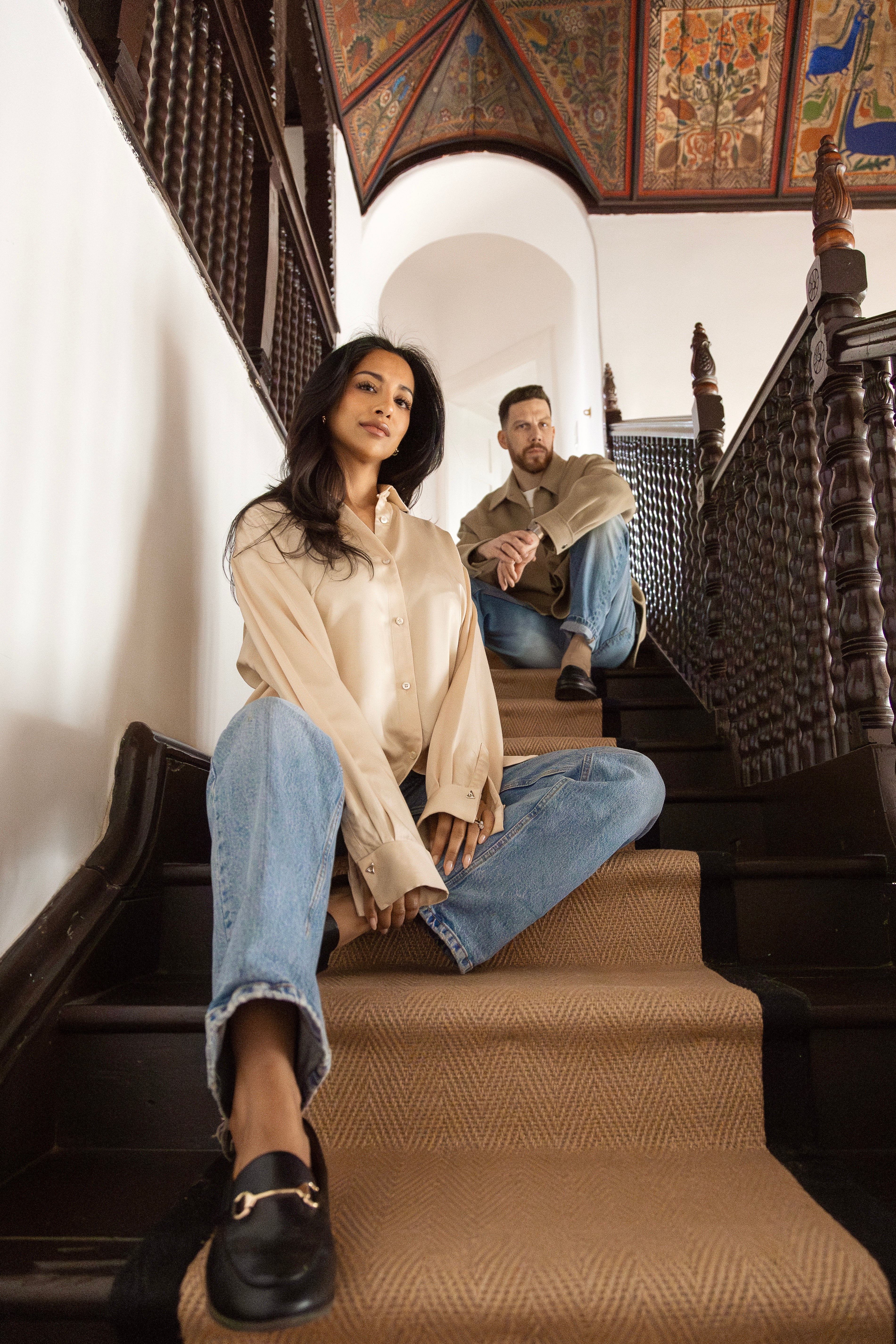Two people sitting on a staircase with ornate wooden railings and a hand painted decorative ceiling.